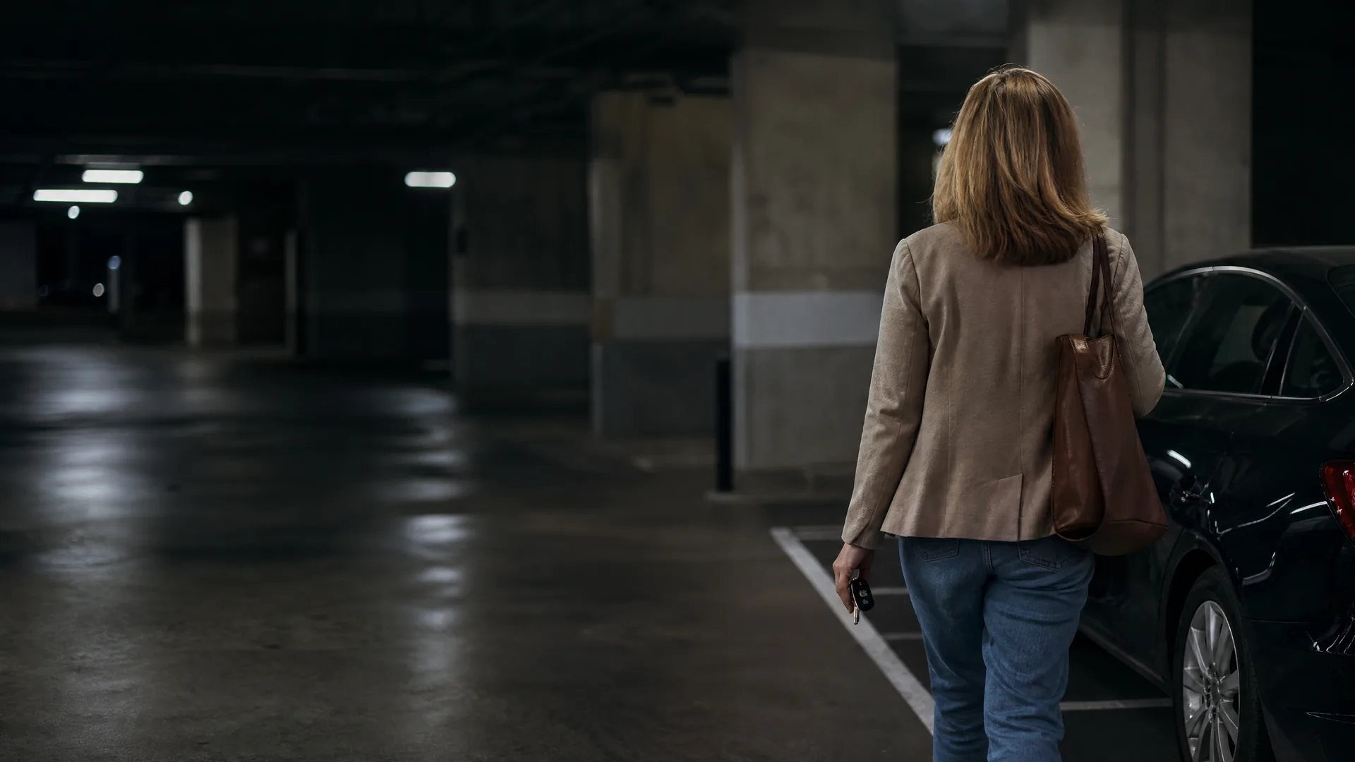 A woman in a tan jacket and jeans with a brown shoulder bag walks away from the camera holding a key fob in a dimly lit underground parking garage beside a dark car.